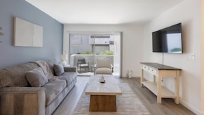 A modern living room with a sofa, coffee table, TV, and a balcony with outdoor furniture. - Landing | Spectacular 1BD, Courtyard (Paradise)
