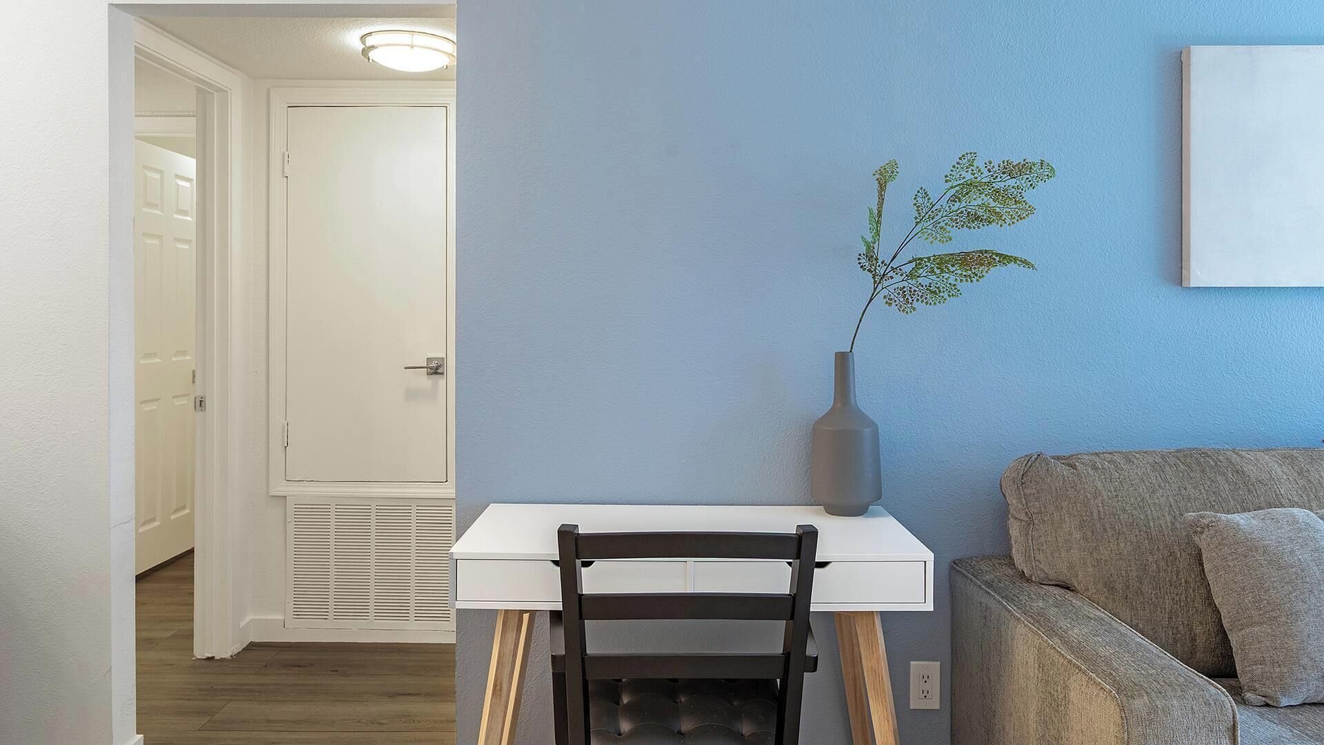 A room with a white desk, a chair, a grey vase with a plant, a grey couch, and a white door.