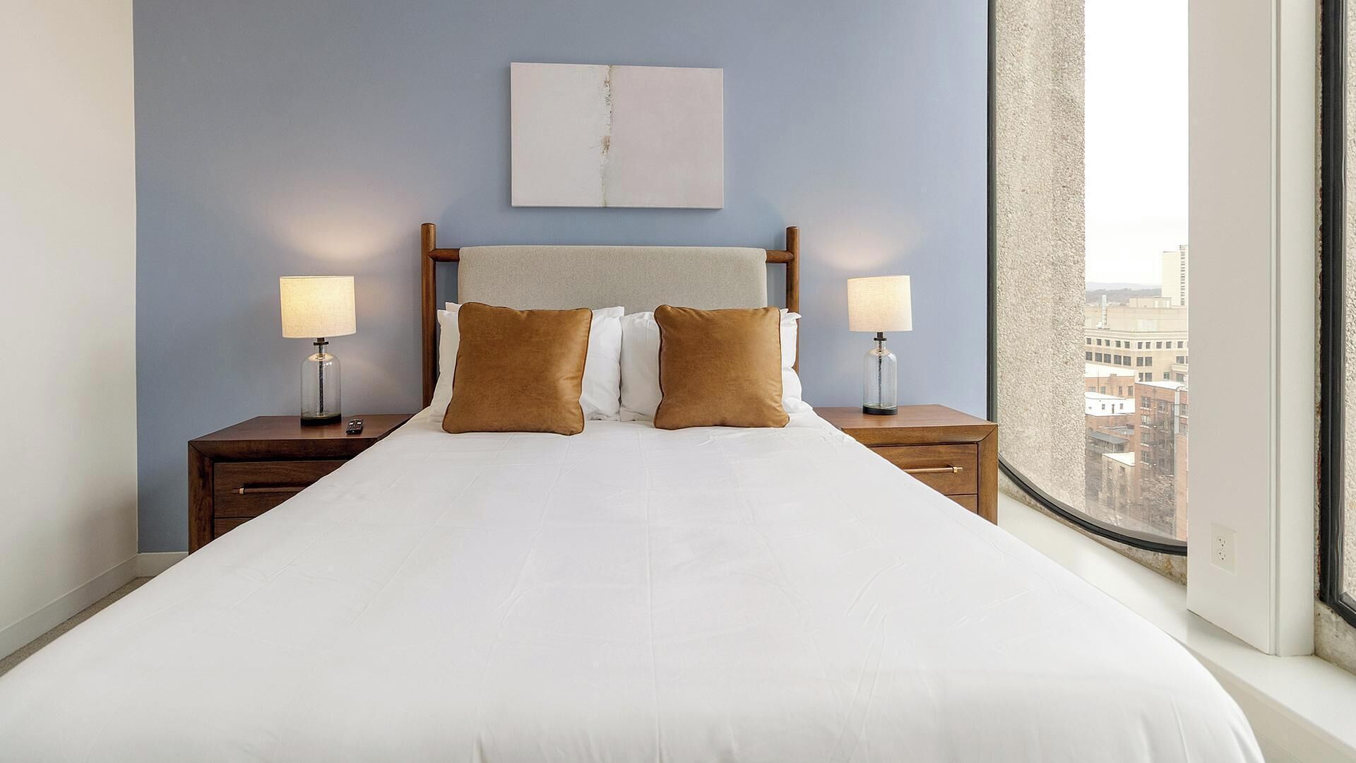 Une chambre d’hôtel avec un grand lit, deux tables de chevet avec des lampes, une tête de lit et une vue sur la ville par une grande fenêtre.