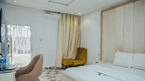 A hotel room with a bed, a desk, a chair, a window with curtains, and an air conditioning unit. - The Rock Aparthotel (Zaria)