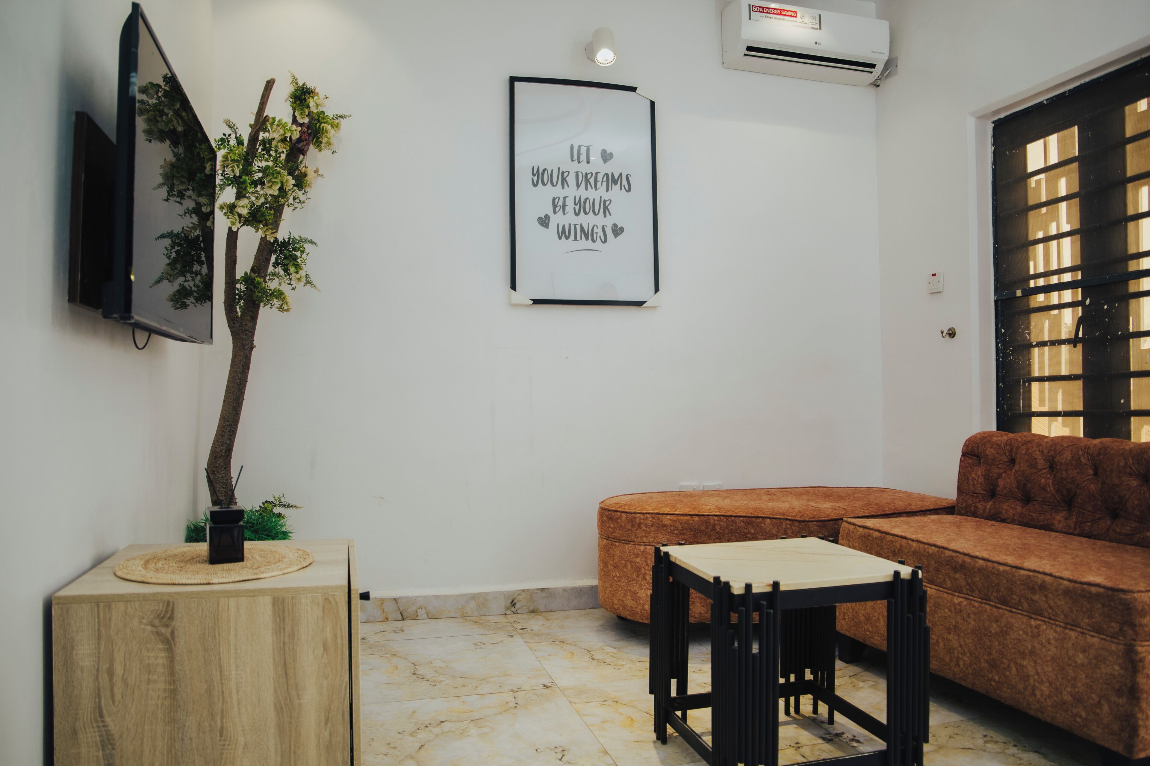 A living room with a sofa, a small table, a mounted TV, a framed inspirational quote, and a potted plant.