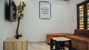 A living room with a sofa, a small table, a mounted TV, a framed inspirational quote, and a potted plant.