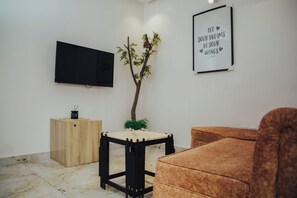 Een moderne woonkamer met een flatscreen televisie, een klein houten kastje, een ronde tafel, een bruine bank, een decoratieve plant en een motiverende quote op de muur.