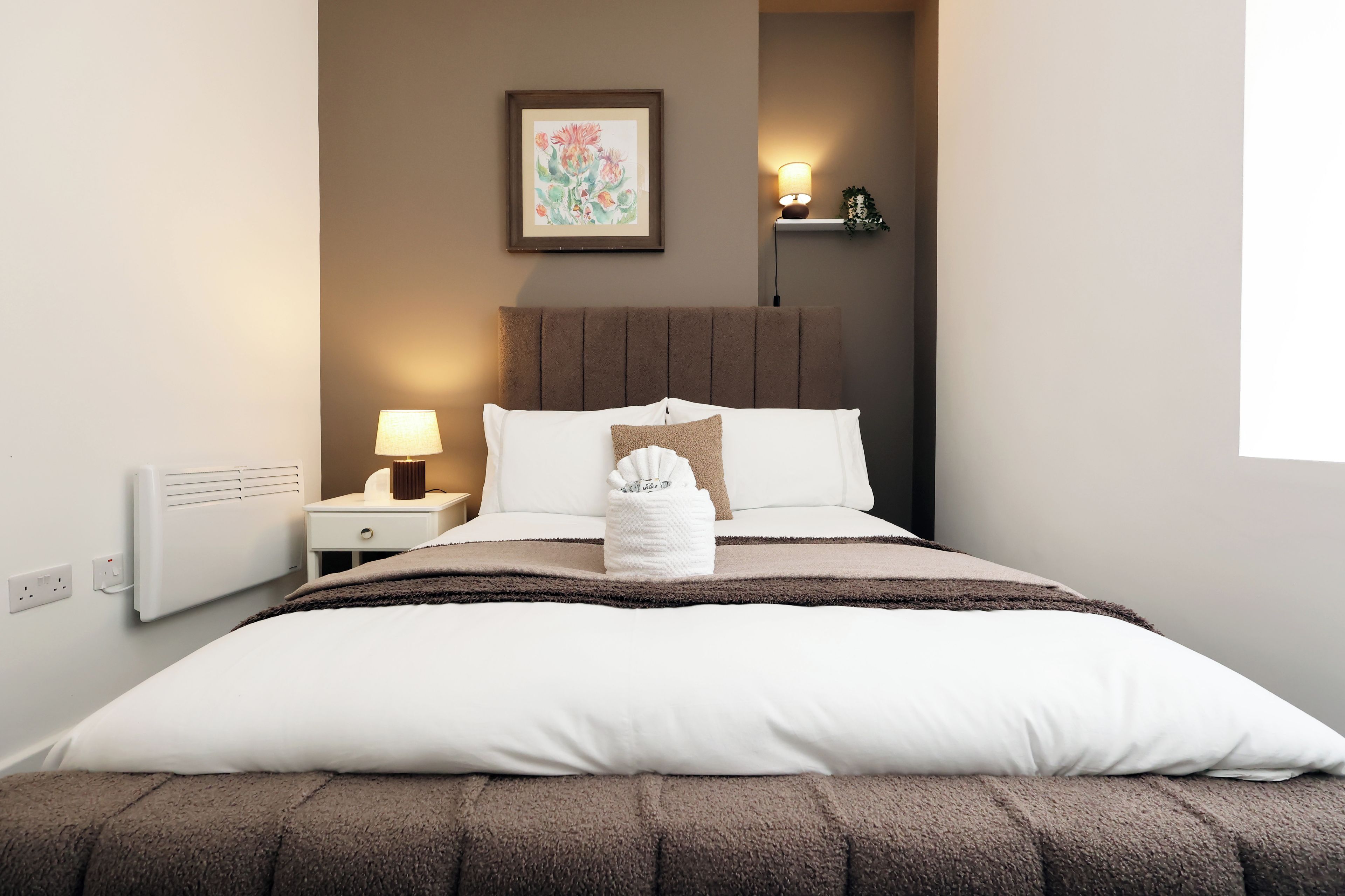 A neatly made bed with a white duvet and brown throw, a bedside lamp, and a wall-mounted picture.