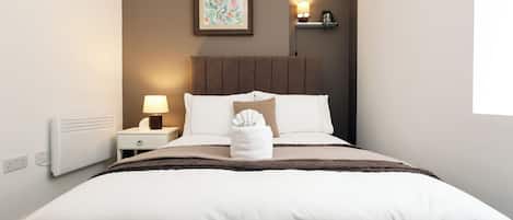 A neatly made bed with a white duvet and brown throw, a bedside lamp, and a wall-mounted picture.