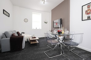 Ein modernes Wohnzimmer mit einem grauen Sofa, einem Glastisch mit Stühlen, einem Fernseher und einem gerahmten Bild an der Wand.