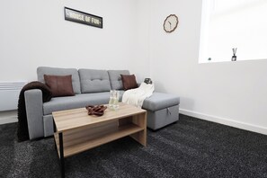 Ein modernes Wohnzimmer mit einem grauen Sofa, einem Holztisch und einem kleinen Beistelltisch mit einem Spielzeugauto und Flaschen.
