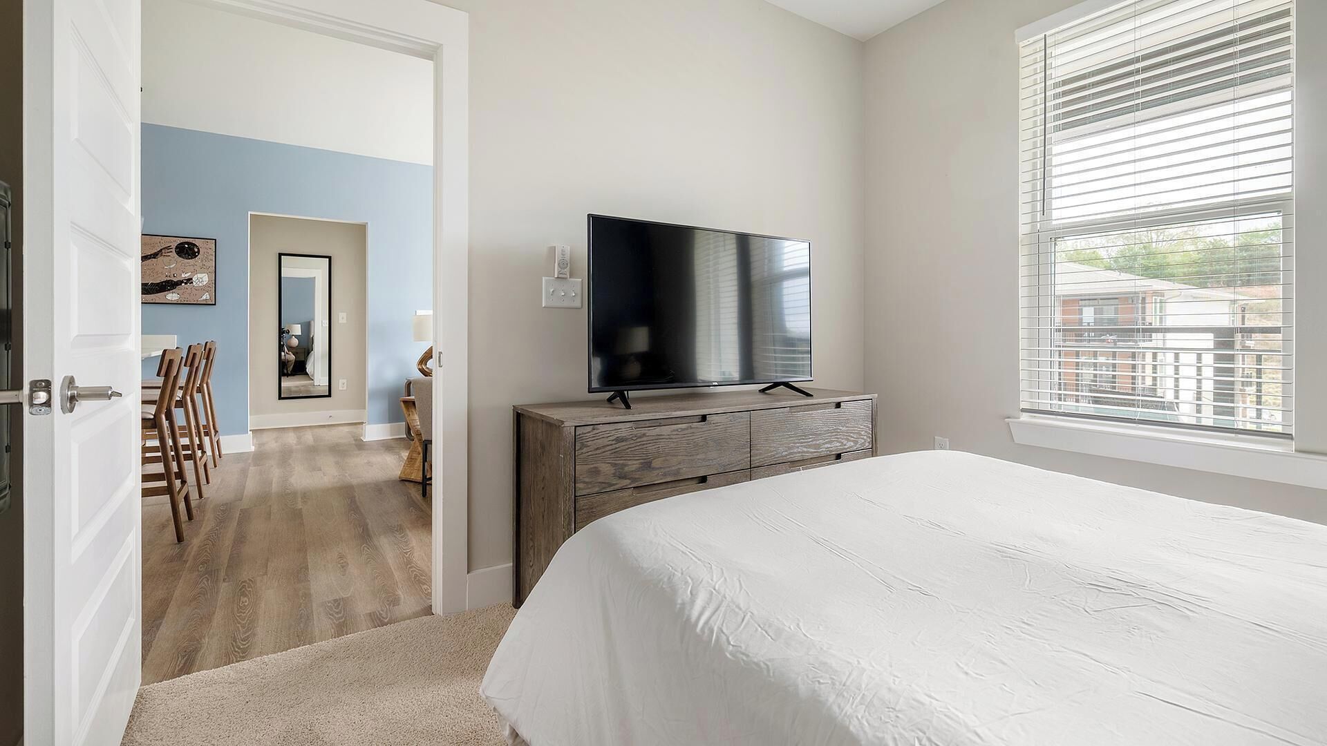 A bedroom with a bed, a TV on a dresser, a window with blinds, and a door leading to another room.