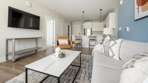 A modern living room with a flat-screen TV, a coffee table, a sofa, and a kitchen in the background. - Landing | Spectacular 2BD, Yoga Studio, Gym (Hickory)