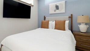 A neatly made bed with white bedding and two brown pillows, a wooden nightstand with a lamp, and a flat-screen TV mounted on the wall. - Landing | Spectacular Studio, Gym, Clubhouse (Houston)