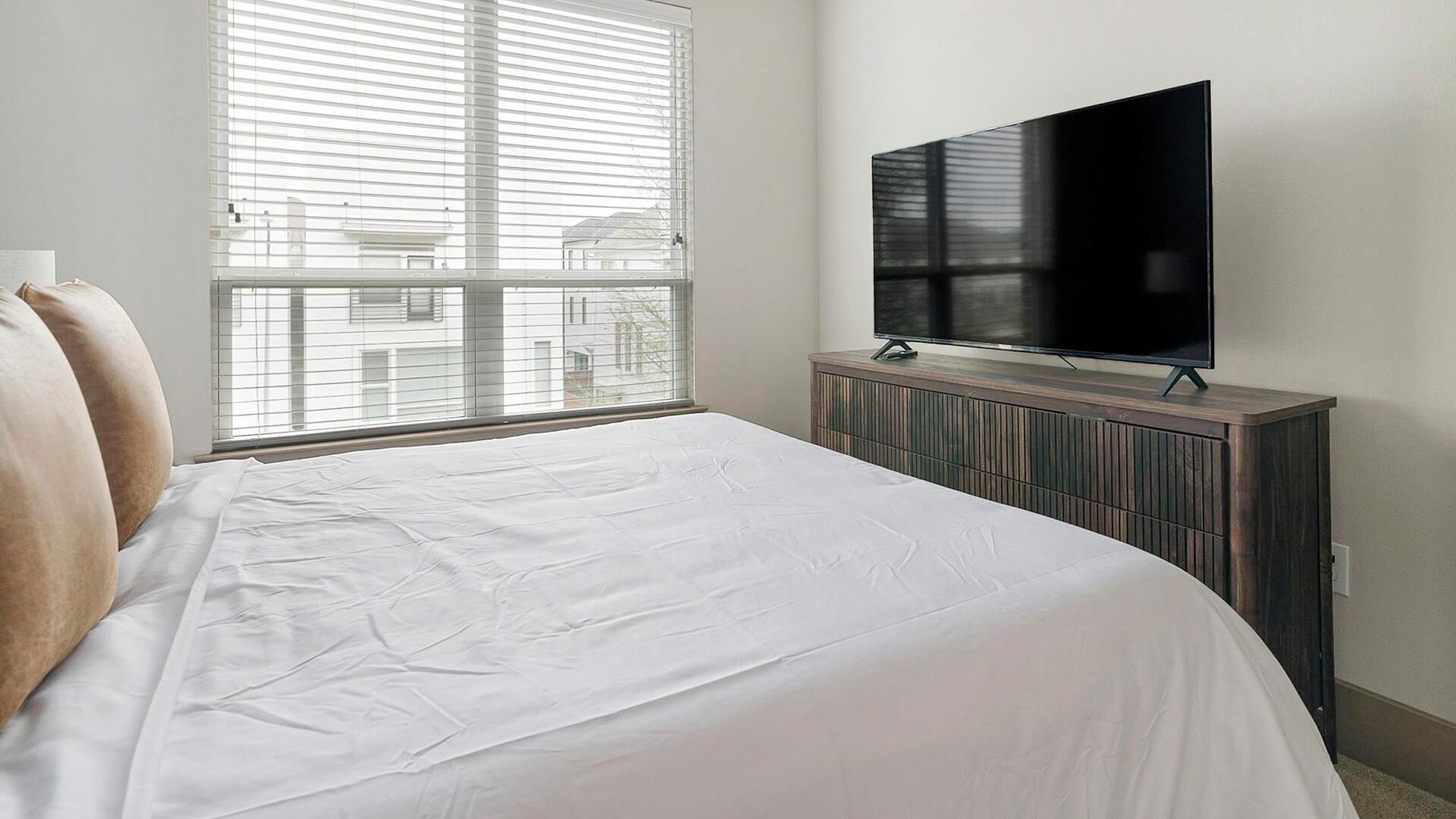 A bedroom with a large bed, a television on a wooden cabinet, and a window with blinds.