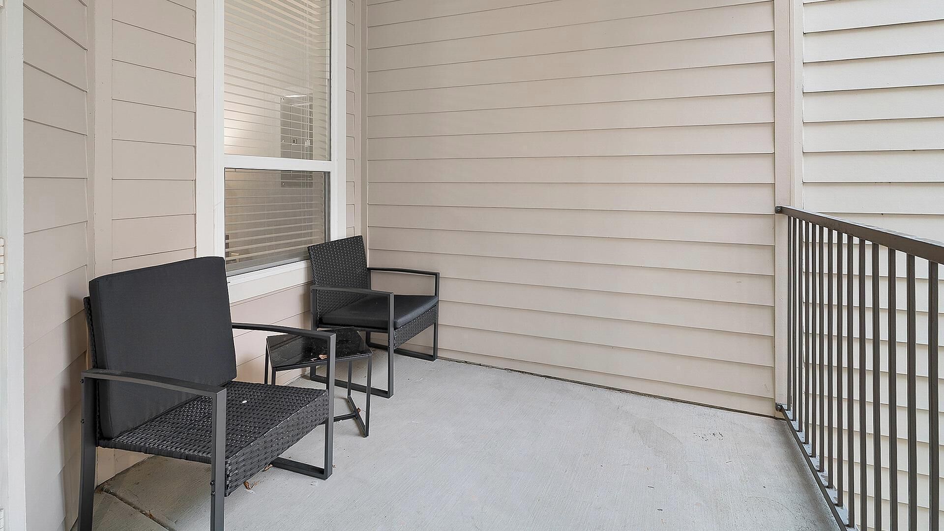 A balcony with two chairs and a table, white siding walls, and a metal railing.
