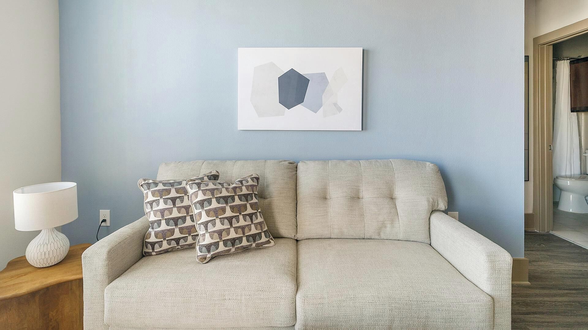 Un canapé beige agrémenté de coussins à motifs, une lampe blanche et une œuvre d’art géométrique accrochée au mur, le tout dans une pièce aux murs bleu clair.