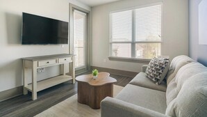 Een woonkamer met een flatscreen televisie, een houten bureau, een ronde salontafel, een grijze bank en grote ramen met zonwering.