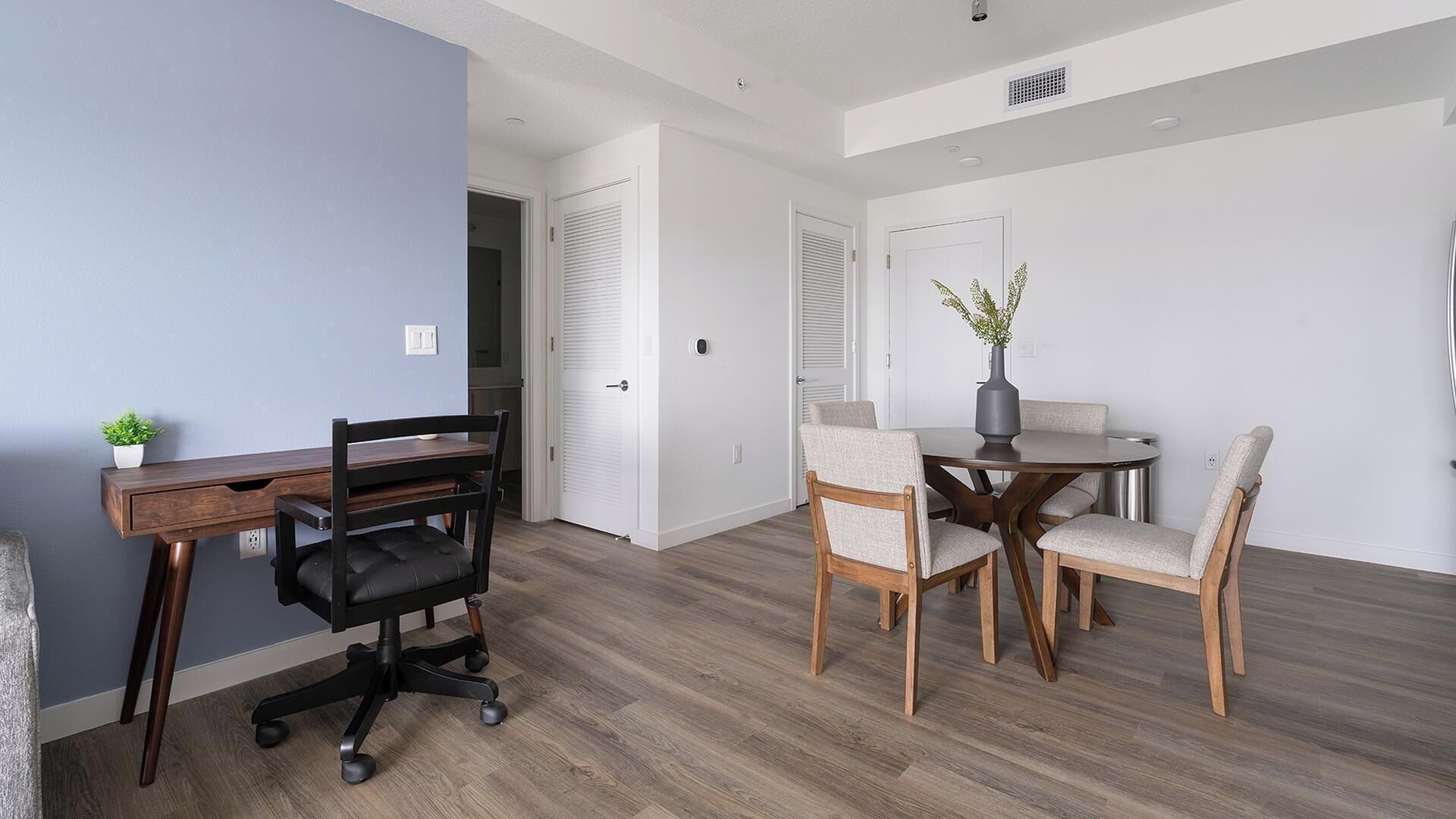 Un appartement moderne comprenant un bureau en bois, une chaise de bureau noire, une table à manger avec quatre chaises et une plante en pot.