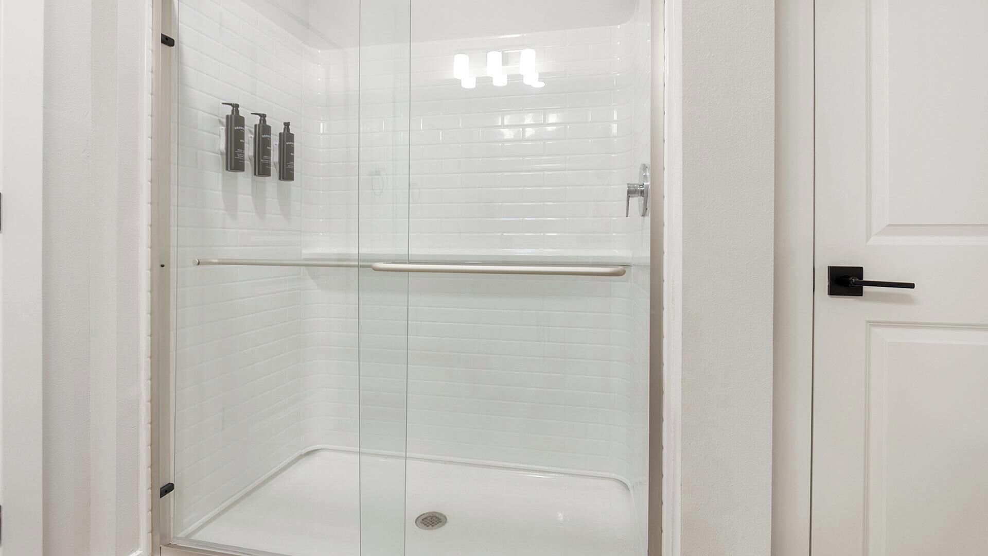 Eine weiße Duschkabine mit Glastür, einer Haltestange und drei Shampooflaschen an der Wand.