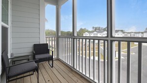 A covered porch with two chairs, a view of a parking lot, and a row of white buildings in the background. - Landing | Stylish 1BD in Shell Point (Port Royal)
