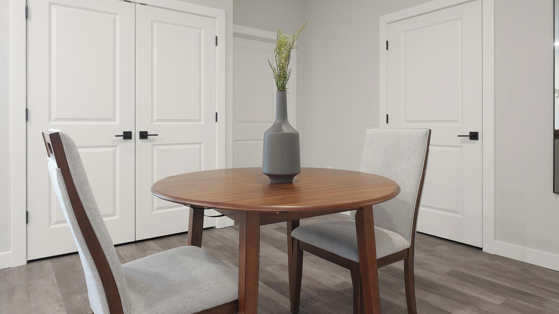 A dining area with a round wooden table, two grey upholstered chairs, and a grey vase with a plant on the table.