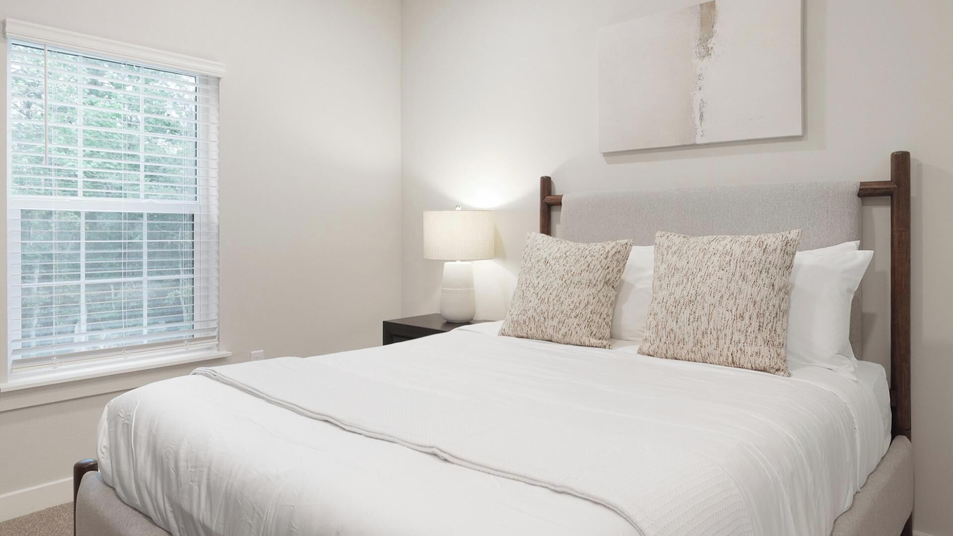 Une chambre avec un grand lit, une table de chevet avec une lampe, une fenêtre avec des stores et un tableau au mur.