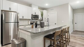 A modern kitchen with stainless steel appliances, white cabinetry, a granite countertop, and a small island with bar stools. - Landing | Stunning 2BD, Gym (Port Royal)
