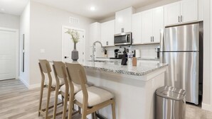 A modern kitchen with white cabinets, a stainless steel refrigerator, a microwave, and a granite countertop. There are three wooden chairs with cushions. - Landing | Modern 2BD in Shell Point (Port Royal)