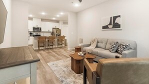 Ein modernes Wohnzimmer mit einer Couch, einem Couchtisch und Sesseln. Im Hintergrund befindet sich eine Küchenzeile mit Schränken und Geräten.