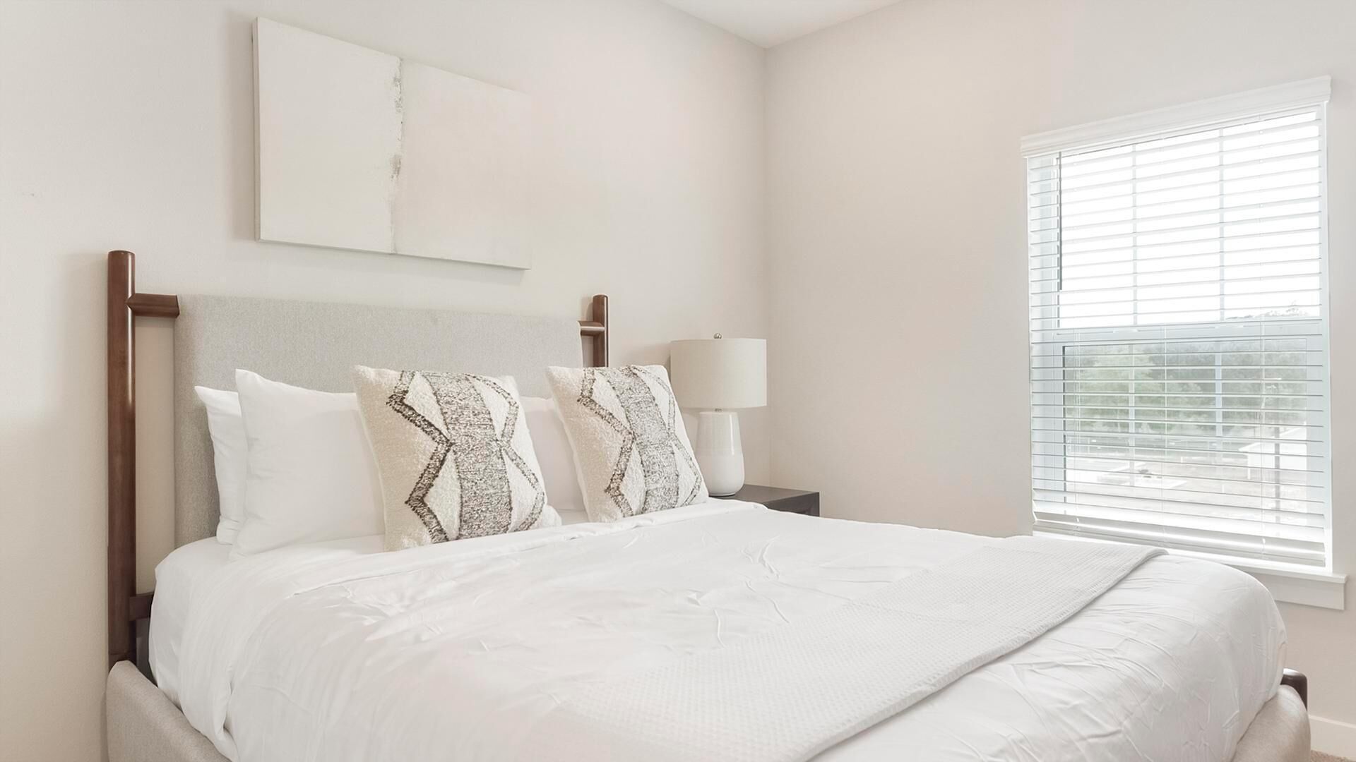 A neatly made bed with a white comforter and two patterned pillows, a bedside table with a lamp, and a window with blinds.