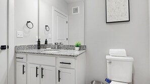 Een badkamer met een wit toilet, een wastafel met granieten blad, een kleine potplant en een kunstwerk in een lijst aan de muur.