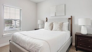 Een slaapkamer met een groot bed, twee nachtkastjes met lampen, een raam met jaloezieën en een schilderij aan de muur.