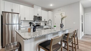 Uma cozinha moderna com armários brancos, eletrodomésticos de aço inoxidável, bancada de granito e uma pequena ilha com banquetas.