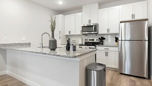 Uma cozinha moderna com armários brancos, eletrodomésticos de aço inoxidável, bancada de granito e uma pequena lixeira.