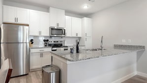 A modern kitchen with white cabinets, stainless steel appliances, granite countertops, and a stainless steel trash can. - Landing | Stunning 1BD in Shell Point (Port Royal)
