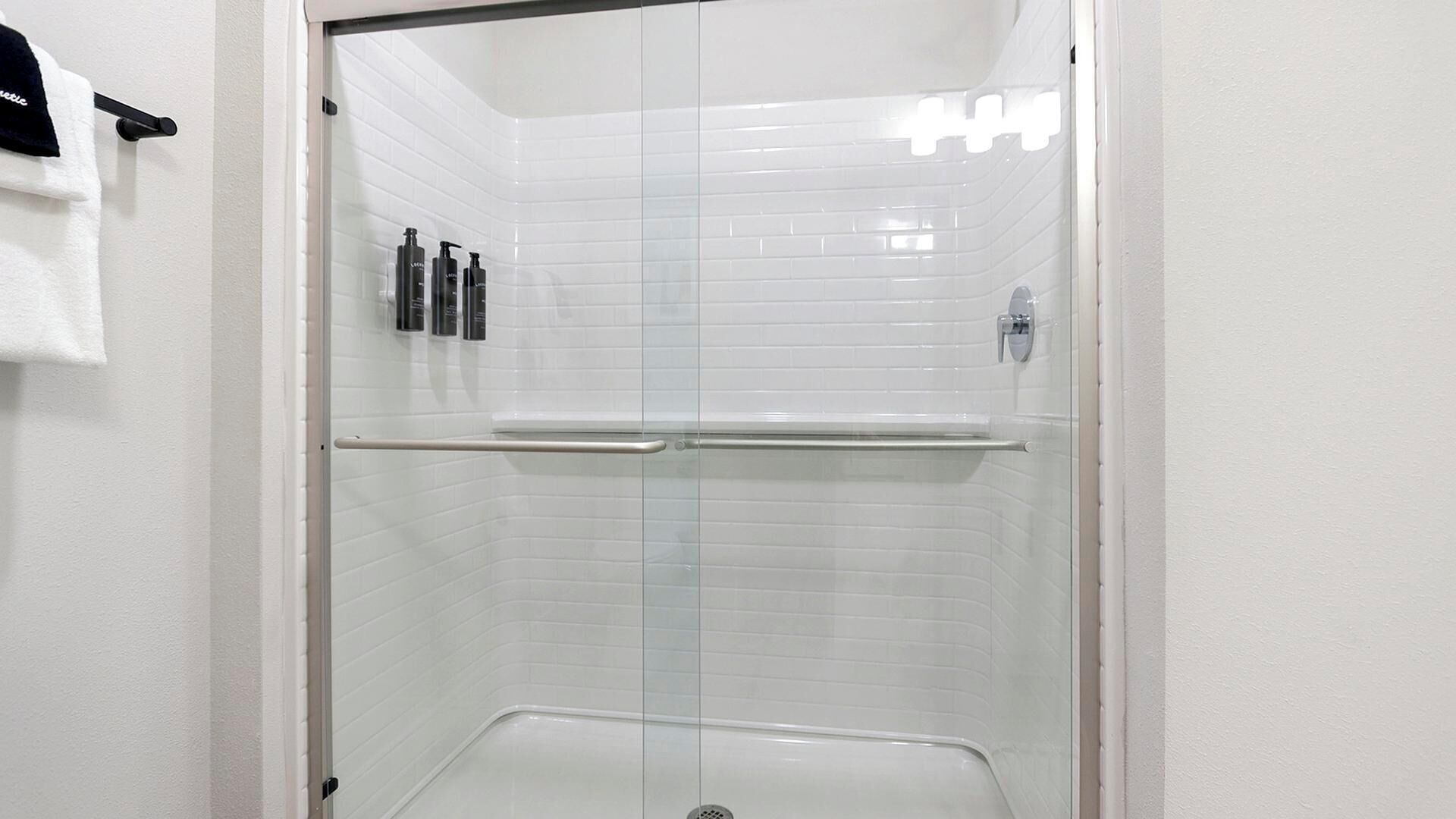 Une cabine de douche fermée par des parois en verre, avec des murs recouverts de carreaux blancs, une barre d’appui en métal et une serviette blanche suspendue à un crochet.
