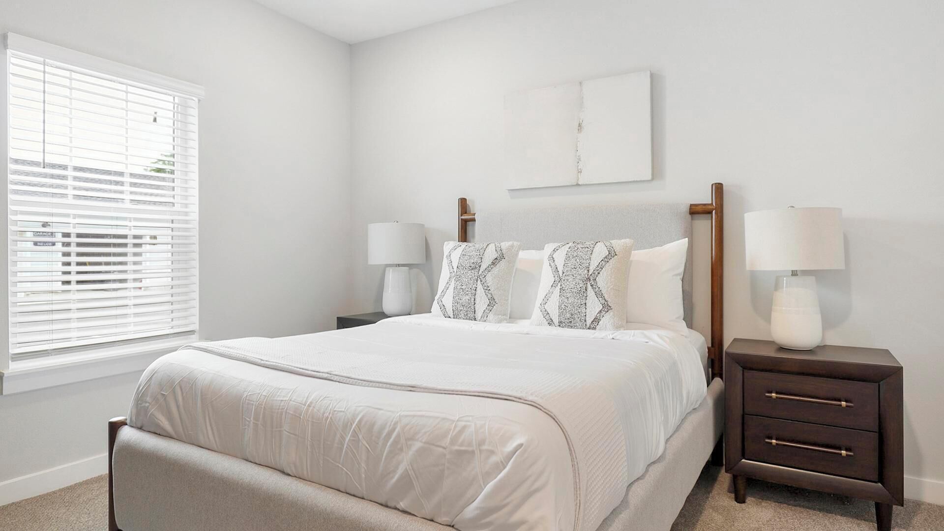 A bedroom with a bed, two bedside tables with lamps, a window with blinds, and a wall-mounted artwork.