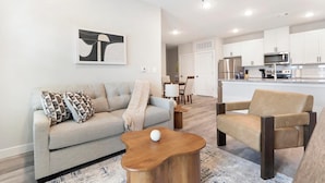 Ein modernes Wohnzimmer mit Sofa, Sessel und Couchtisch, einem Essbereich und einer Küche mit weißen Schränken und Edelstahlgeräten.