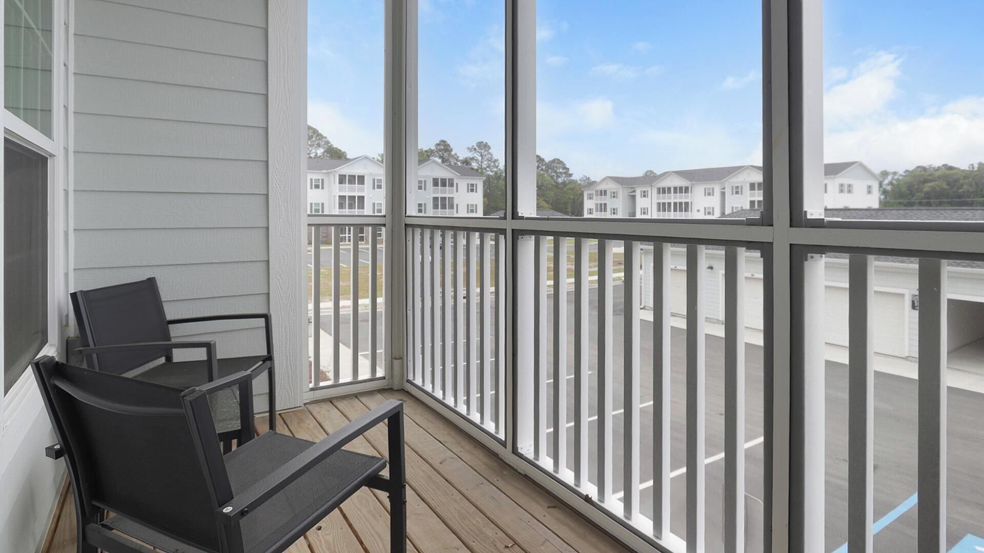 A balcony with a view of a row of white houses, a wooden deck, and two black chairs.