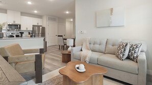 Un salon moderne comprenant un canapé, un fauteuil et une table basse, une cuisine équipée d’appareils électroménagers en acier inoxydable et un coin repas avec une table et des chaises.