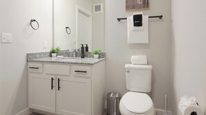 A bathroom with a white toilet, a sink with a granite countertop, a mirror, and a towel rack with a towel. - Landing | Beautiful 1BD in Shell Point (Port Royal)