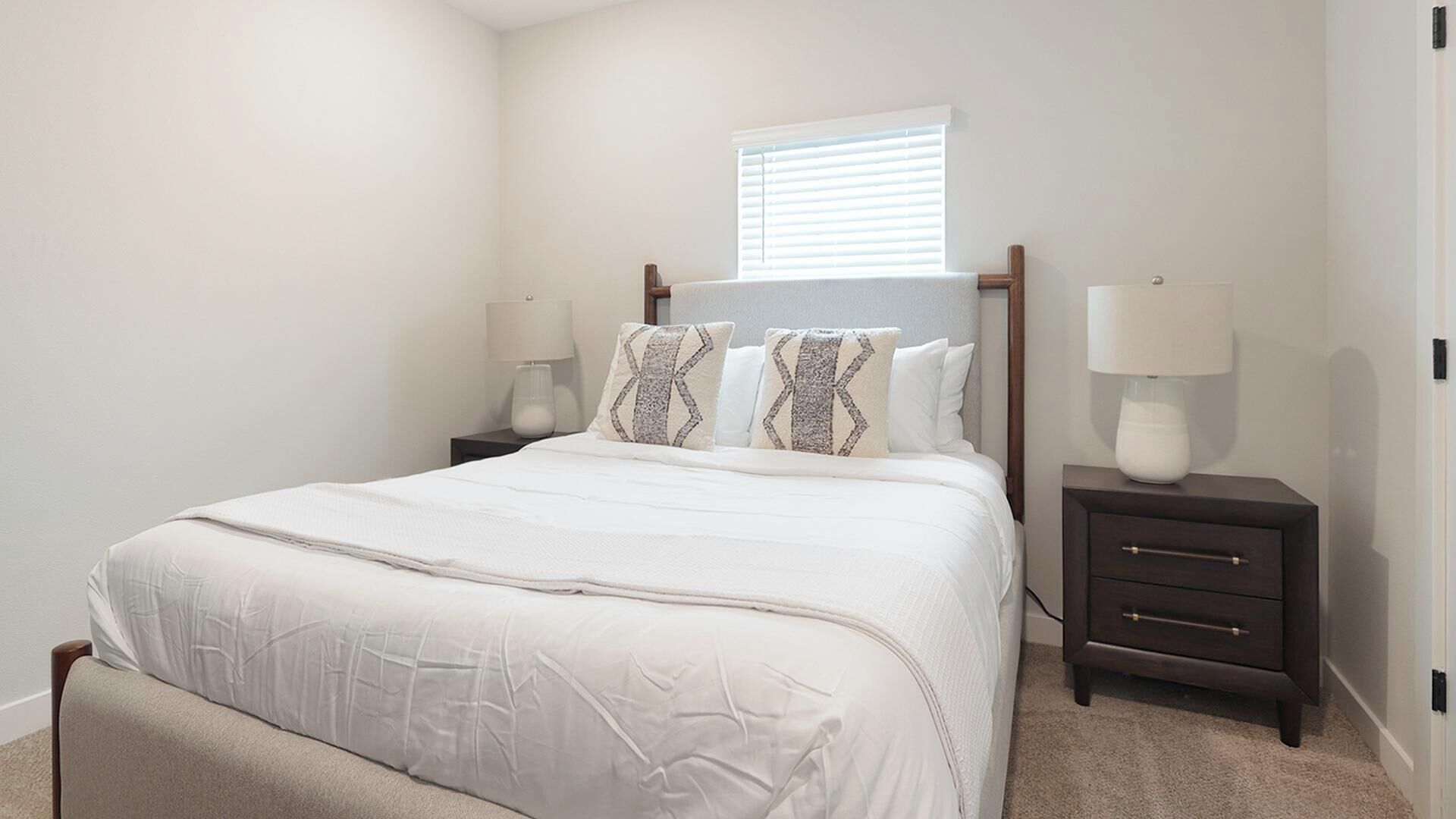 A bedroom with a bed, two bedside tables, a window with blinds, and a nightstand with a lamp.