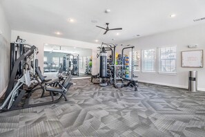 Uma academia com diversos equipamentos de exercício, um ventilador de teto e vista para o exterior através de amplas janelas.
