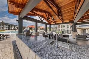 A covered outdoor bar area with a granite countertop, seating, and a pool in the background. - Landing | Stylish 2BD in Medical Center (San Antonio)