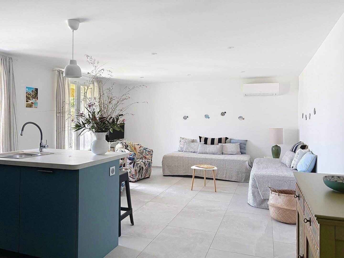A modern kitchen with a central island, a dining area with a sofa and a small table, and a bedroom with a bed and a bedside table.