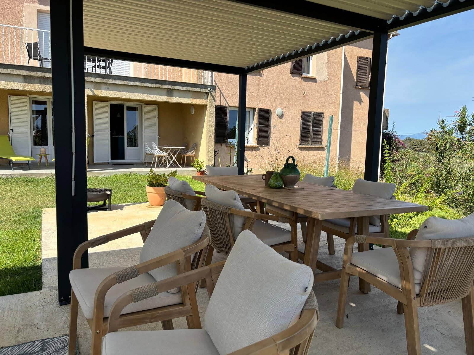 Eine Terrasse im Freien mit einem Holztisch, Korbstühlen, einem überdachten Bereich und Blick auf ein Gebäude mit Fenstern und einer Tür.