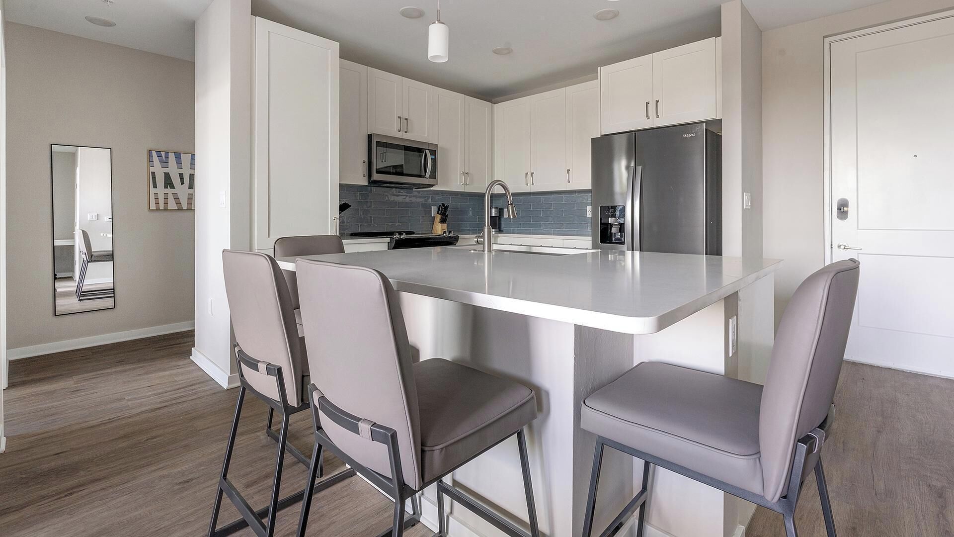 A modern kitchen with white cabinets, a stainless steel refrigerator, a microwave, and a sink. There are two bar stools and a large island with a sink.