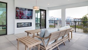 A modern balcony with a wooden table and sofa, large windows, and a mounted TV. - Landing | Modern 2BD, Clubhouse, Yoga Studio (Bonita Springs)