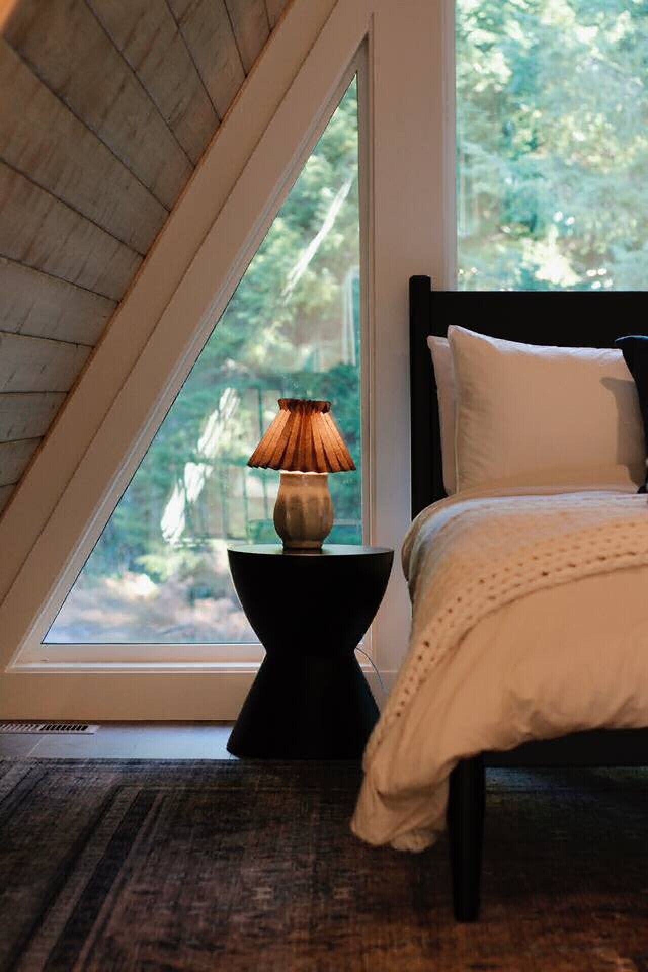 A bedroom with a sloped ceiling, a bed with white bedding, a black nightstand with a lamp, and a view of greenery outside.