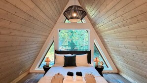 A bedroom with a sloped ceiling, a bed with pillows, a bedside lamp, and a window with a view of greenery. - NEW Mt Rainier Aframe | Sauna+Hot Tub | White Pass (Packwood)