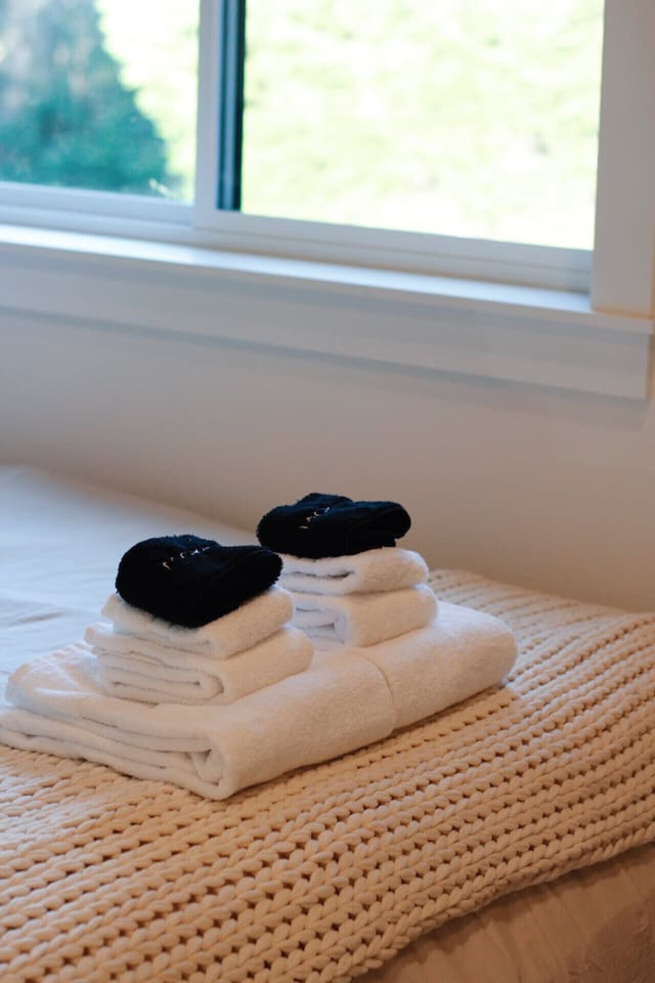 En prydligt vikt hög med vita handdukar ligger på en beige, vävd matta, placerad på en säng nära ett fönster med utsikt över grönskan utanför.