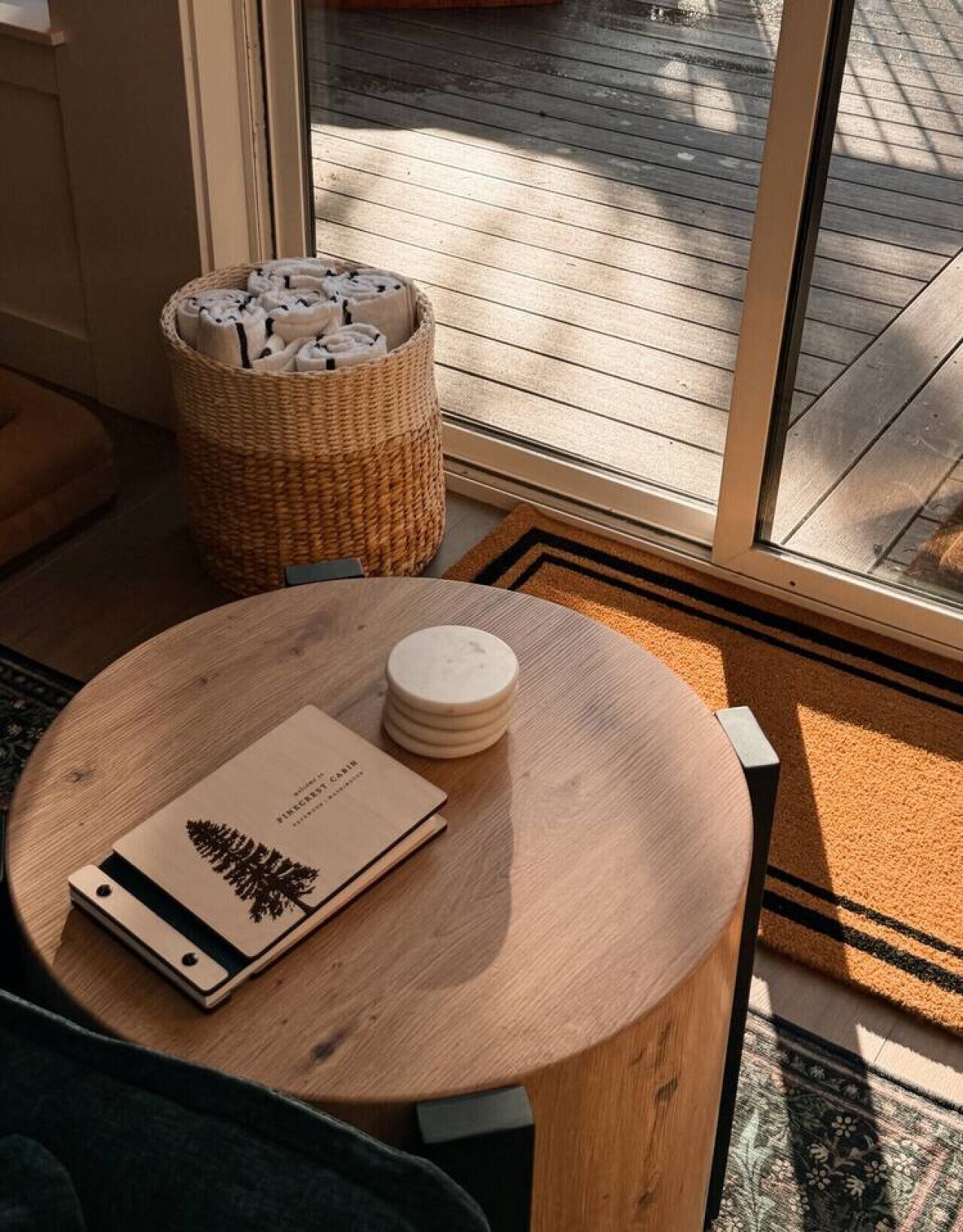 A wooden table with a book and coasters, a wicker basket, and a view of a wooden deck through a glass door.