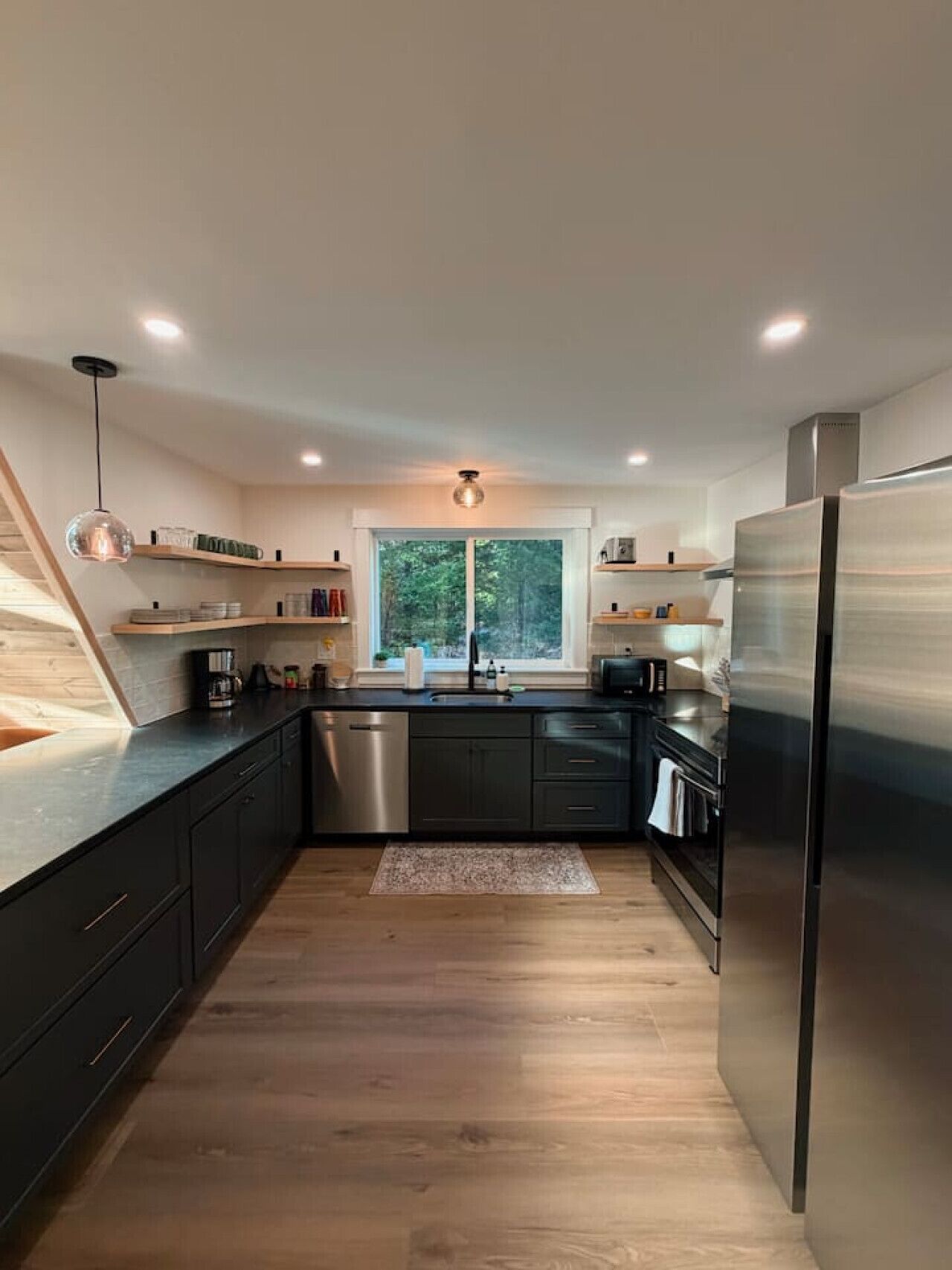 A modern kitchen with dark cabinetry, a stainless steel refrigerator, and a window with a view of trees.